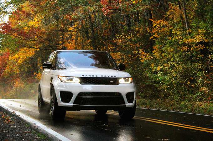 Range Rover Sport driving on road by woods during fall front side view.