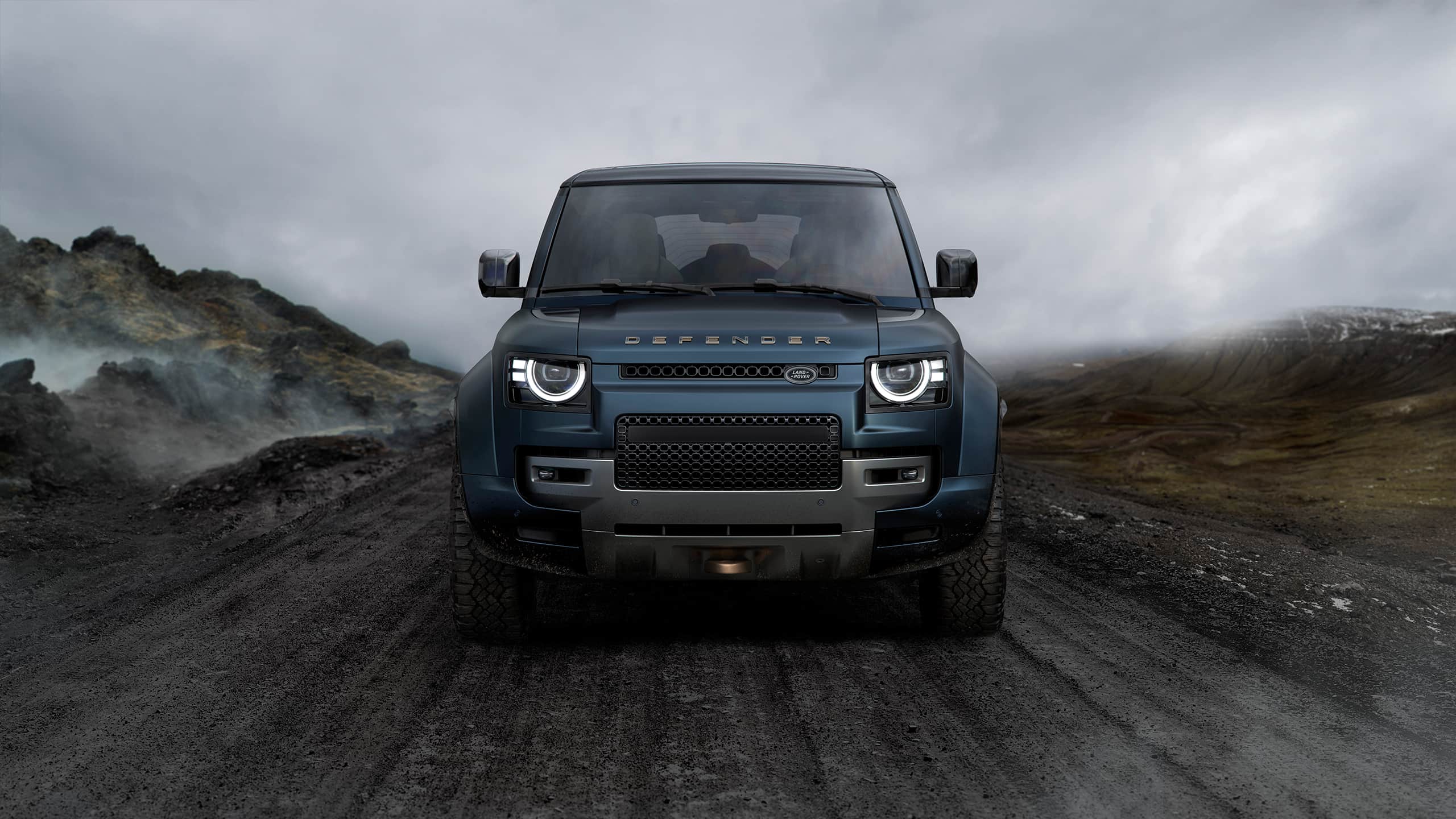 Front view of a blue Defender on a rugged mountain road under cloudy skies.